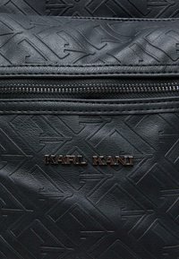 Sac en cuir noir avec motif en relief, doté d'une fermeture éclair et d'un logo métallique "KARL KANI" à l'avant. Texture lisse.