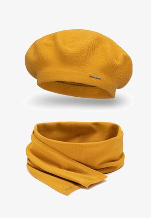 Mustár sárga kötött beret sapka texturált mintával és kis fém logóval. Hozzáillő kötött sál sima felülettel és körbetekerős stílussal.