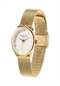 Montre à bracelet en maille dorée avec un cadran rond blanc, présentant des index horaires dorés, le logo Victoria Hyde, et un fermoir gravé "Acier inoxydable".