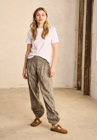 Femme debout à l'intérieur portant un t-shirt blanc, un pantalon ample à motifs et des chaussures marron sans lacets, devant un mur texturé uni.