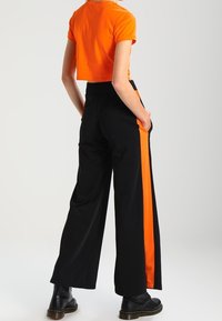 Persona vestida con pantalones anchos negros con franjas laterales de color naranja brillante, una camiseta corta naranja y botas negras, de pie con las manos en los bolsillos.