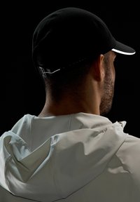 Gorra negra con acentos reflectantes, forma redondeada y visera blanca. Se lleva con una chaqueta con capucha de color claro que presenta una textura en la tela.