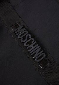 Czarna torba tekstylna z teksturowanym, wypukłym logo "MOSCHINO" na pasku. Powierzchnia jest gładka z widocznymi szwami.