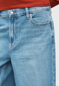 Κοντινό πλάνο ανοιχτό μπλε τζιν τζιν με μπροστινή τσέπη και μεταλλικό κουμπί, φορεμένο με κόκκινο ριγωτό τοπ.