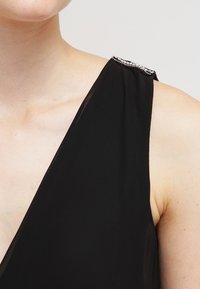 Czarna sukienka bez rękawów z głębokim dekoltem w kształcie V, lekkiego materiału oraz ozdobnym detalem na ramionach z srebrnymi akcentami.
