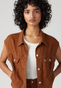 Colete de denim castanho sem mangas com botões prateados e bolsos no peito, combinado com uma t-shirt branca de malha. Textura suave, design casual.