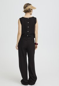 Femme portant un haut noir sans manches avec boutons dans le dos et un pantalon à jambes larges, un chapeau visière beige, un bracelet rouge et un bracelet argenté, debout devant un fond blanc.