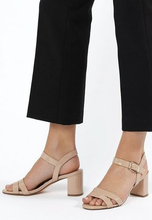 Voeten met nudekleurige open teen sandalen met blokhak en enkelbandjes, gecombineerd met zwarte cropped broeken op een witte achtergrond.