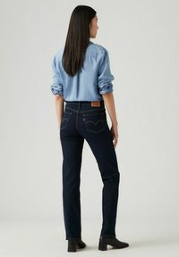 Vaqueros azul oscuro con dos bolsillos traseros y emblema de Levi's, combinados con una camisa azul clara, mostrados desde atrás. Se ven zapatos de tacón negros.