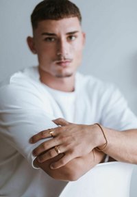 Camicia bianca a maniche corte, mani incrociate con tre accessori dorati: un anello, un bracciale, un anello decorato. Sfondo neutro.