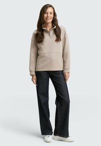 Beige, langelzaam pullover met een kwart rits, hoge kraag en zachte textuur, gecombineerd met donkere wijde jeans en witte sneakers.