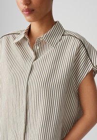Blusa a righe in tessuto crema con sottili linee verticali marroni. Colletto classico, maniche corte con una vestibilità comoda e patta con bottoni.
