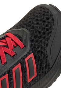 Zapatilla deportiva negra con parte superior de malla texturizada, cordones rojos y acentos a rayas. Suela de goma suave con un diseño de dibujo para mejor agarre.