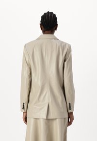 Heled helebeige nahkblazer, millel on lõikega krae, kohandatud istuvus ja ventilaatoritaotlus seljas. Omab musti nööbidetailide aksessuaare varrukatel.