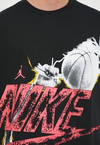 Mustra musta puuvillane T-särk, millel on silmapaistev roosa "NIKE" kiri, korvpalluri siluett ja abstraktsed valged ja kollased graafilised elemendid.