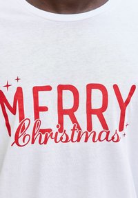 Bílá bavlněná tričko s nápisem "VESKRZE Vánoce" v tučných červených písmenech. Text obsahuje ozdobné prvky jako hvězdy.