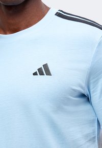 Κοντινό πλάνο ενός ατόμου που φοράει ελαφρύ γαλάζιο πουκάμισο Adidas με μαύρες ρίγες στον ώμο και το λογότυπο Adidas στο στήθος.
