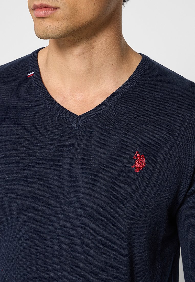 Dunkelblaues V-Ausschnitt-Pullover aus Strickstoff, mit einem roten gestickten Logo auf der linken Brust und einem kleinen rot-weißen Akzent am Kragen.