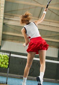 Tennisspelare i en vit och röd outfit serverar på en bana, med en korg med klara gröna tennisbollar i närheten. Tyget verkar vara lätt.