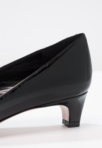 Chaussure en cuir verni noir avec un petit talon, un bout pointu et une texture lisse. Présente une couture discrète le long du bord d'ouverture.