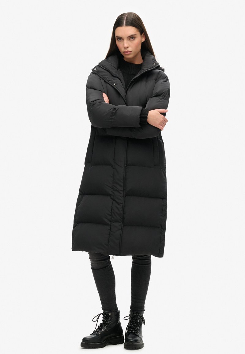 Zwart pufferjack met een lange silhouet, voorzien van een ritsluiting, grote zakken en een capuchon. Het materiaal heeft een gladde, matte afwerking.