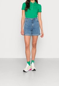 Polo vert avec boutons blancs, short en denim taille haute avec ourlets retroussés, et baskets blanches avec accents roses et chaussettes vertes.