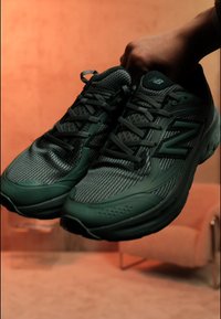 Άτομο κρατάει ένα ζευγάρι σκούρα πράσινα αθλητικά παπούτσια New Balance με λεπτομέρειες από δίχτυ και κορδόνια, με φόντο ένα θολό εσωτερικό.