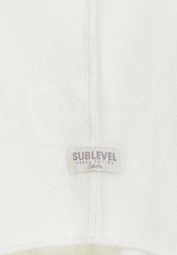 Weißer textiles Stoff mit einem genähten Etikett, auf dem "SUBLEVEL URBAN EDITION Kollektion" in helllila und grauen Tönen steht.