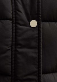 Czarna pikowana tkanina z przeszytymi sekcjami, z okrągłym, jasnym zatrzaskiem w pobliżu krawędzi, zapewniająca gładką i miękką teksturę.