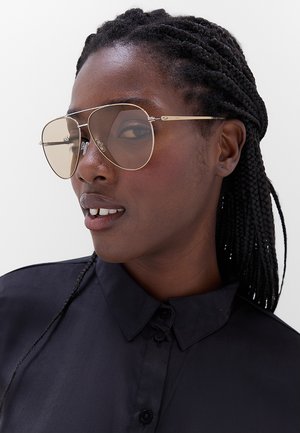 Donna con i capelli intrecciati che indossa grandi occhiali da aviatore con montatura dorata e una camicia nera con colletto, che guarda leggermente di lato con un'espressione neutra.