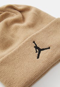 Bež pleteni beanie sa crnim izvezenim logotipom osobe koja izvodi visoki udarac na preklopnom rubu.
