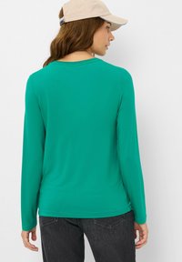 T-shirt à manches longues, ajusté, en tissu turquoise vif, avec une texture lisse et un col rond. Porté avec un jean foncé, le modèle porte une casquette beige.