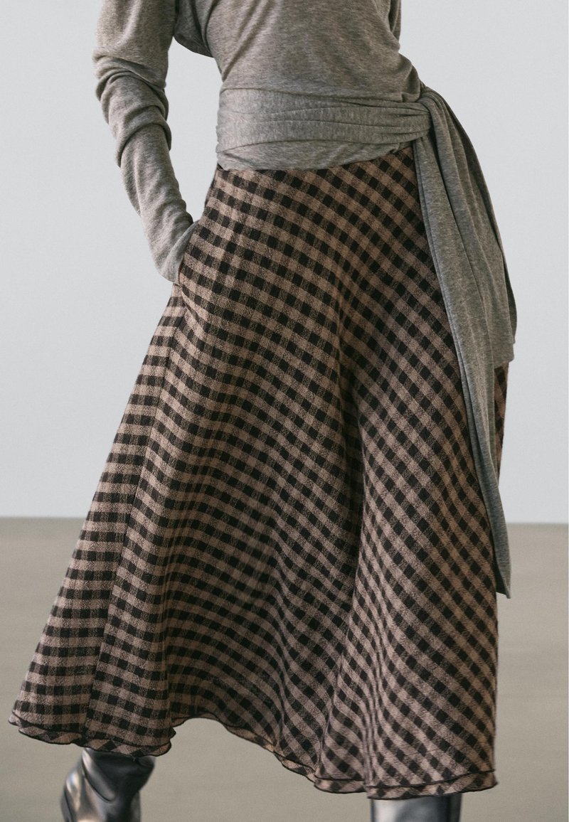 Karierter Rock mit schwarzem und braunem Muster, fließendem Stoff und gezacktem Saum. Kombiniert mit einem grauen Langarmshirt.