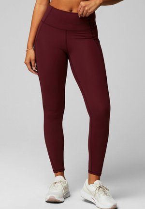 Burgundy high-waisted leggings made of stretchy fabric with flatlock seams, smooth texture, and a fitted silhouette, paired with white sneakers.