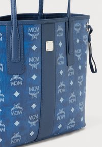 Sac fourre-tout bleu avec des accents en cuir et un motif embossé arborant le logo MCM. Comprend deux poignées pour l'épaule et une plaque de marque en métal.