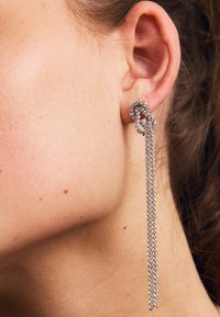 Garas sudraba auskari ar mezglu dizainu, kuros ir piekārti ķēdes elementi. Virsma ir ar teksturētu apdari un nelieliem, iegrieztiem detaļām.