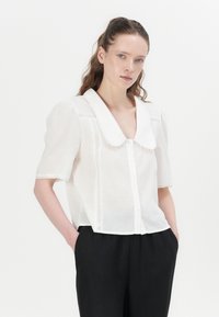 Hvit kortermet bluse med stor krage, med knappelukking og blonder detaljer. Kombinert med svarte høyt midje bukser.
