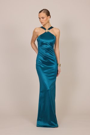 Femeie care poartă o rochie de seară din satin teal, cu guler halter, broșă aurie în formă de fluture, cercei aurii și o brățară aurie, pozând pe un fundal bej.