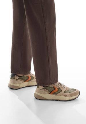 Sneakers beige e verde oliva con accenti arancioni, materiali in camoscio e rete, suola spessa e dettagli testurizzati; indossati con pantaloni marroni.