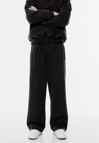 Čierne tepláky vyrobené z mäkkej látky, s elastickým pásom na stiahnutie, širokými nohavicami a bočnými vreckami. Doplnia ich biele tenisky.