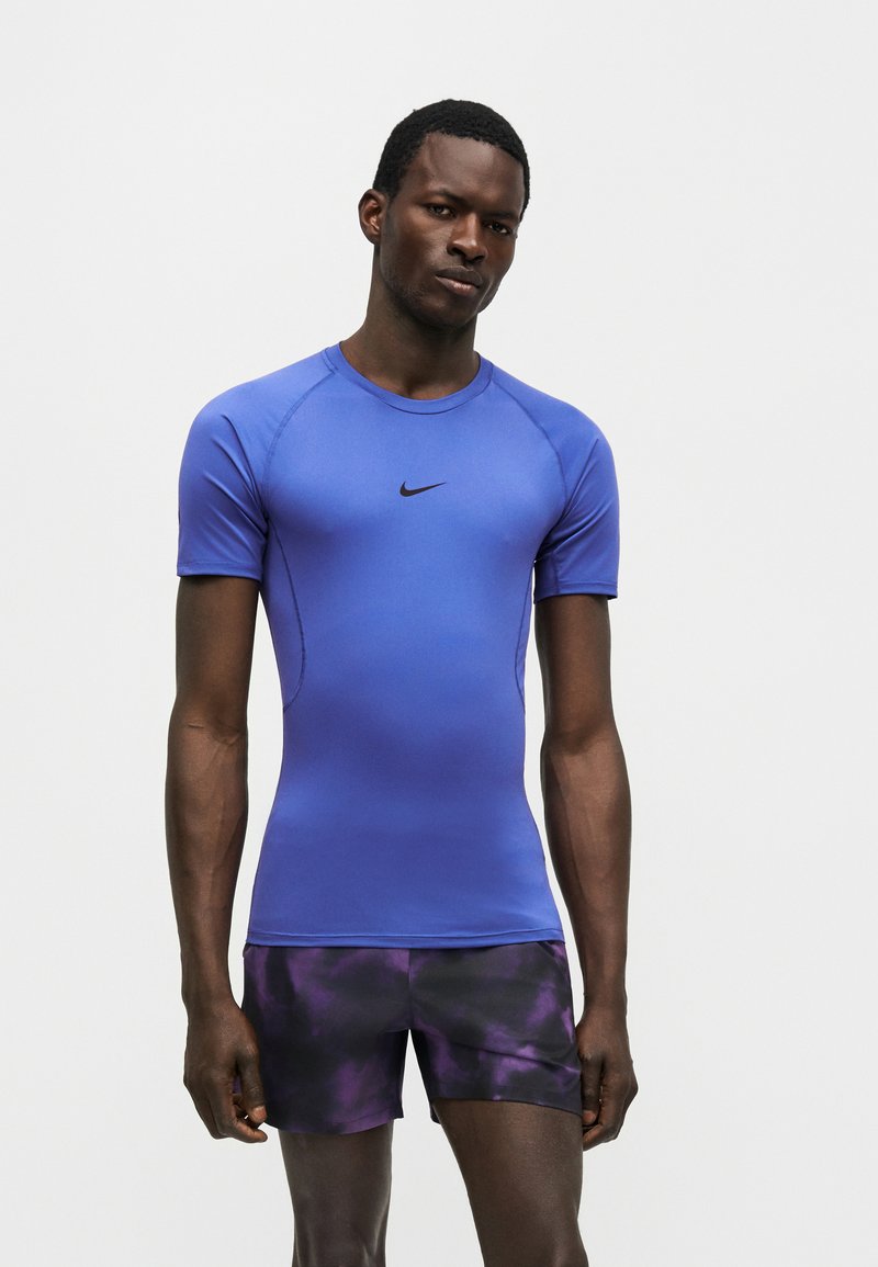 Atlétický muž nosí priliehavé modré krátke tričko značky Nike a čierno-fialové vzorované šortky, stojí na jednoduchom bielom pozadí.