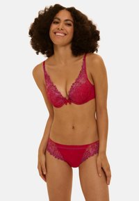 Frau mit lockigem Haar lächelnd, trägt roten Spitzen-BH und passende Unterwäsche, steht vor einem schlichten, hellen Hintergrund.