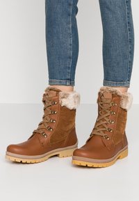 Brune snørebukser med en overdel i semsket skinn, faux pelskant, gummisåle og metallisk hardwaredetaljer. Standard ankelhøyde.