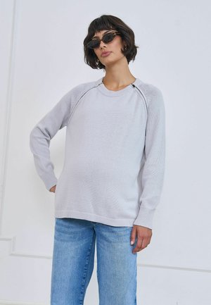 Femme aux cheveux courts et foncés portant des lunettes de soleil, un pull gris clair avec des détails de fermeture éclair et un jean bleu, debout contre un mur blanc.