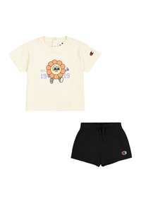 T-shirt in cotone crema con grafica di un fiore da cartone animato, pantaloni shorts elastici neri con patch logo, entrambi progettati per un abbigliamento casual.