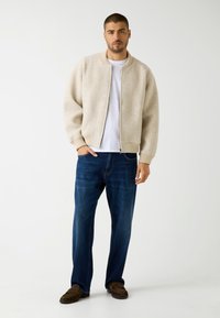 Beige wollen bomberjack met geribbelde kraag en manchetten, gedragen over een wit t-shirt, gecombineerd met donkerblauwe jeans en bruine mocassinschoenen.