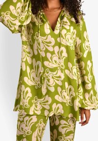 Top verde a fantasia floreale con maniche a campana e scollatura a V. Il materiale appare leggero con una texture morbida. Sono visibili pantaloni floreali coordinati.