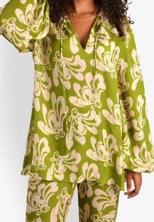 Grün gemusterte Bluse mit Glockenärmeln und V-Ausschnitt. Das Material sieht leicht und weich aus. Dazu passende florale Hosen sind sichtbar.