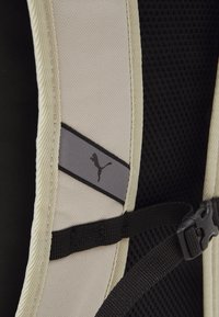 Sac à dos noir et beige avec une texture en maille, un patch de logo, des bretelles ajustables et une fermeture à boucle. Tissu durable avec des coutures renforcées.