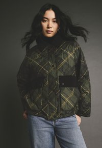 Jachetă verde matlasată cu un model de carouri galbene, având un guler negru, nasturi tip snap și două buzunare frontale. Purtați-o peste blugi albaștri.